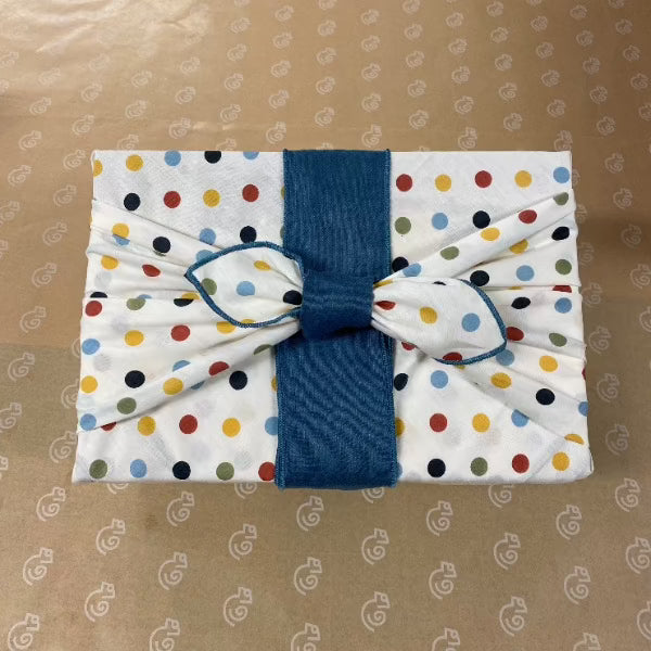 This Furoshiki or fabric gift wrap has cute spots and dots on a light cream background. The earthy multicoloured spots are red, yellow, green, blue, and black. They are solid colours and all the same size dots. This particular picture has a blue Belly Band on our large fabric gift wrap. It gives the knot a distinctive bow style. It is a sophisticated look but still fun and can celebrate any occasion.