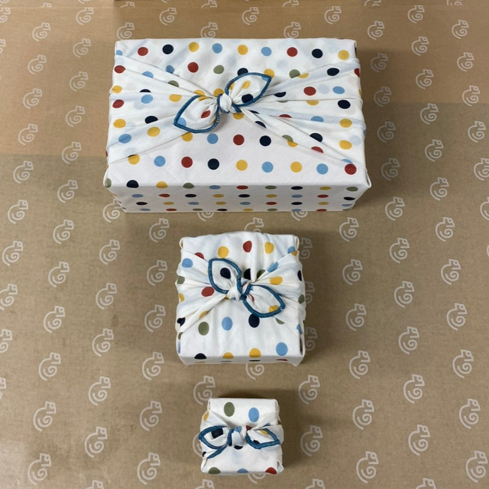 This Furoshiki or fabric gift wrap has cute spots and dots on a light cream background. The earthy multicoloured spots are red, yellow, green, blue, and black. They are solid colours and all the same size dots. This picture displays 3 sizes, extra small, small , and medium.