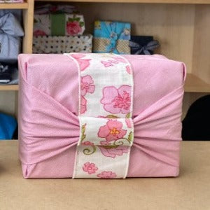 The fabric gift wrap in a rectangle shape with a neat bow ties on the top. The fabric is of a tiny pink pinstripe that almost looks like one colour. Displayed in a mid soft pink  with white edging. There is a band of vintage floral fabric that goes under the knotted bow and makes the gift extra special