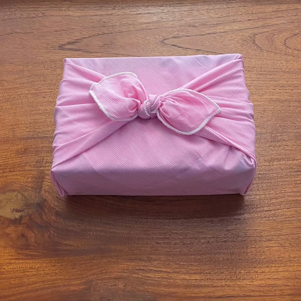 The fabric gift wrap in a rectangle shape with a neat bow ties on the top. The fabric is of a tiny pink pinstripe that almost looks like one colour. Displayed in a mid soft pink  with white edging